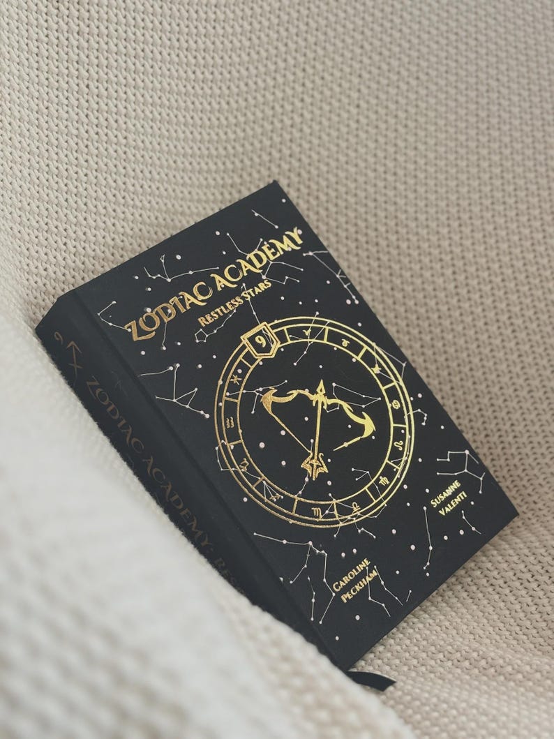 May include: A black hardback book titled Zodiac Academy: Restless Stars, featuring gold foil constellations and a central bow and arrow symbol. The cover displays the number 9 and the authors' names, Caroline Peckham and Susanne Valenti.