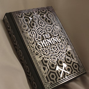 Peut inclure: Un livre à couverture rigide intitulé "The Shining" avec un motif géométrique argenté et noir. La couverture présente un labyrinthe et deux haches croisées. Le livre a un signet en ruban argenté.