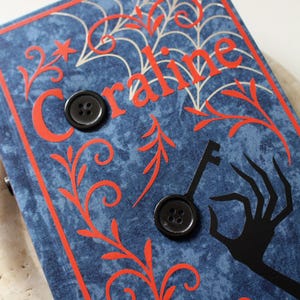 May include: A blue fabric-covered book with the title "Coraline" in red lettering. The cover features a spiderweb design, decorative swirls, and a black key and hand silhouette. Two black buttons are attached to the cover.
