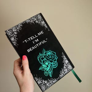 以下が含まれることがあります： 白い花の縁取りと「T-TELL ME I'M BEAUTIFUL」の文字が入った黒いジャーナル。 ターコイズのバラとスカルのデザインが特徴です。 緑のリボンブックマークが含まれています。