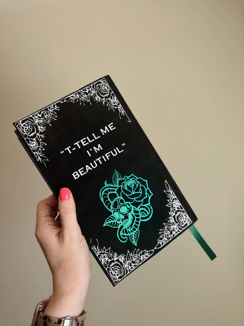 以下が含まれることがあります： 白い花の縁取りと「T-TELL ME I'M BEAUTIFUL」の文字が入った黒いジャーナル。 ターコイズのバラとスカルのデザインが特徴です。 緑のリボンブックマークが含まれています。
