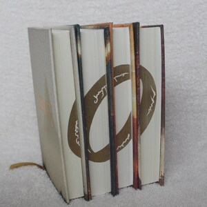 May include: A set of three hardback books standing upright, featuring a continuous gold ring design printed across the white page edges. The book covers are textured in shades of tan, brown, and orange, with a thin gold ribbon bookmark visible.
