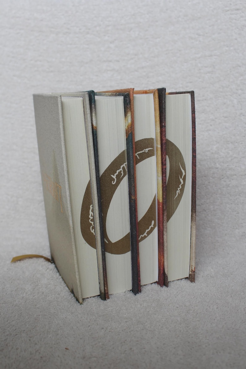 May include: A set of three hardback books standing upright, featuring a continuous gold ring design printed across the white page edges. The book covers are textured in shades of tan, brown, and orange, with a thin gold ribbon bookmark visible.