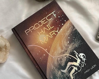 Op de afbeelding: Hardcover boek getiteld "PROJECT HAIL MARY" met een ruimtethema. De omslag toont een astronaut, planeten en sterren in bruin, oranje en blauw. De naam van de auteur, ANDY WEIR, staat onderaan gedrukt.