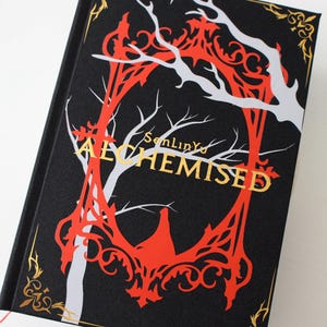 Può includere: Un libro con copertina rigida nera con un disegno di albero rosso e bianco. Il titolo "ALCHEMISED" è in lettere dorate. Accenti dorati decorano gli angoli. Probabilmente un romanzo fantasy.