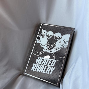 Può includere: Un taccuino grigio scuro con un bordo argentato e le parole "HEATED RIVALRY" in bianco. La copertina presenta una grafica bianca di due giocatori di hockey che si fronteggiano con le loro mazze incrociate.