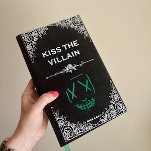 以下が含まれることがあります： 「KISS THE VILLAIN」というタイトルの黒い本。緑色の顔のデザインと白い花のアクセントが特徴です。本には緑色のリボンしおりがあり、著者の名前であるRina Kentが印刷されています。