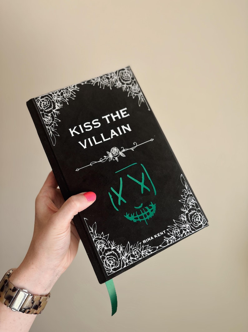 以下が含まれることがあります： 「KISS THE VILLAIN」というタイトルの黒い本。緑色の顔のデザインと白い花のアクセントが特徴です。本には緑色のリボンしおりがあり、著者の名前であるRina Kentが印刷されています。