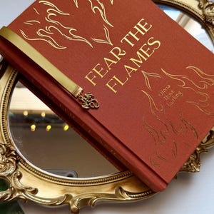 May include: A hardback book titled Fear the Flames by Olivia Rose Darling with a burnt orange fabric cover, gold foil flame illustrations, and a gold ribbon bookmark featuring a small metal dragon charm attached to the end of the ribbon.