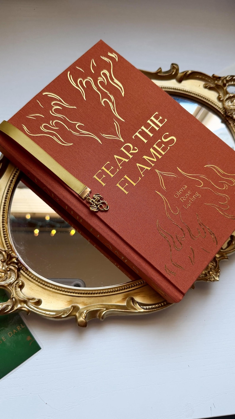 May include: A hardback book titled Fear the Flames by Olivia Rose Darling with a burnt orange fabric cover, gold foil flame illustrations, and a gold ribbon bookmark featuring a small metal dragon charm attached to the end of the ribbon.