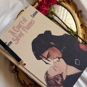 May include: A hardback book titled A Court of Silver Flames with a cream-coloured fabric cover featuring an illustration of two figures kissing. A black ribbon bookmark with a small red bead charm is attached to the book, which rests on a gold tray.