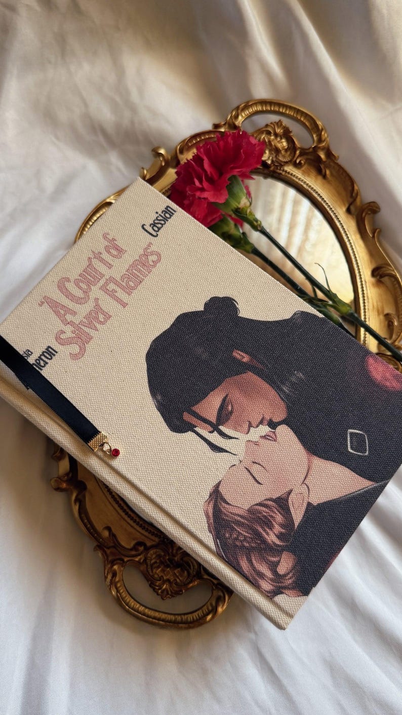 May include: A hardback book titled A Court of Silver Flames with a cream-coloured fabric cover featuring an illustration of two figures kissing. A black ribbon bookmark with a small red bead charm is attached to the book, which rests on a gold tray.