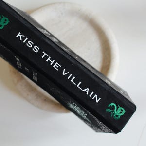 以下が含まれることがあります： 白文字で「KISS THE VILLAIN」と書かれた黒い本。緑色の装飾が施されています。本は明るい色の円形の表面に置かれています。本の背表紙が見え、タイトルが表示されています。
