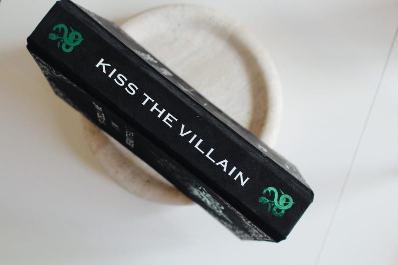 以下が含まれることがあります： 白文字で「KISS THE VILLAIN」と書かれた黒い本。緑色の装飾が施されています。本は明るい色の円形の表面に置かれています。本の背表紙が見え、タイトルが表示されています。