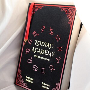 May include: A hardback book titled Zodiac Academy: The Awakening by Caroline Peckham and Susanne Valenti. The black cover features gold lettering, pink zodiac symbols arranged in a circle, decorative pink corner borders, and a red ribbon bookmark.