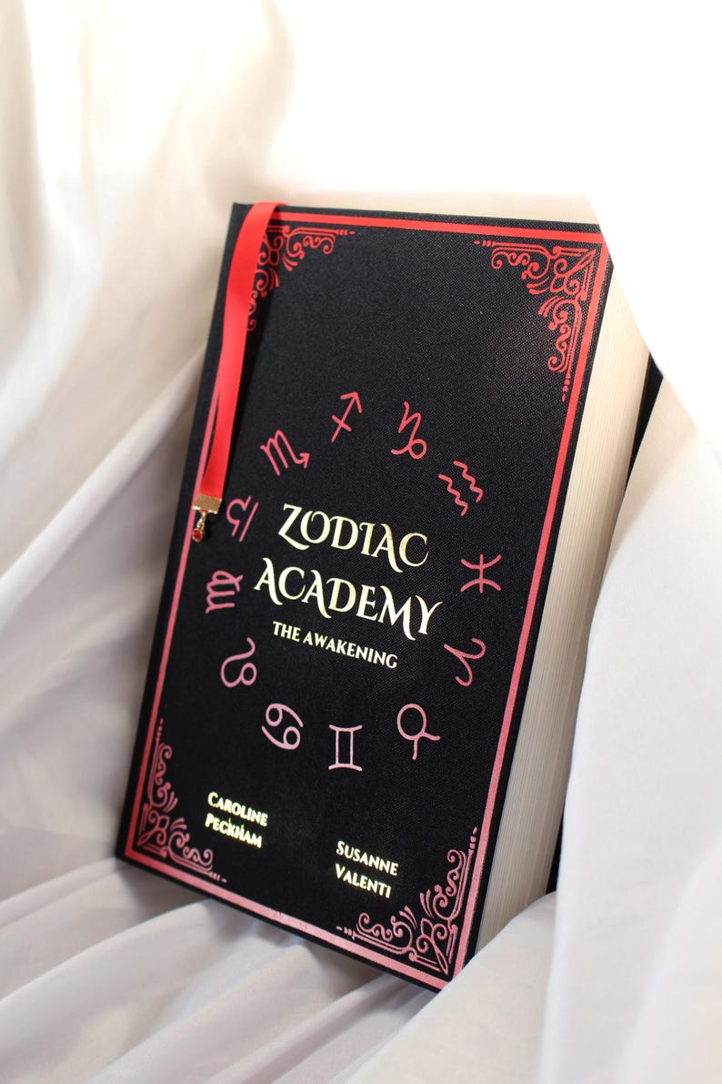 May include: A hardback book titled Zodiac Academy: The Awakening by Caroline Peckham and Susanne Valenti. The black cover features gold lettering, pink zodiac symbols arranged in a circle, decorative pink corner borders, and a red ribbon bookmark.