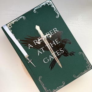 Peut inclure: Livre à couverture rigide intitulé "A Reaper at the Gates" avec une couverture vert foncé. La couverture présente un corbeau noir, une épée argentée et des coins argentés décoratifs. Un signet en ruban blanc avec un charme d'épée est inséré.