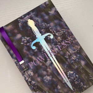 May include: A hardback book titled Powerless by Lauren Roberts featuring a cover image of purple lavender flowers. A large, iridescent, holographic sword graphic is centred on the cover. A purple ribbon bookmark with a small metal sword charm is attached.