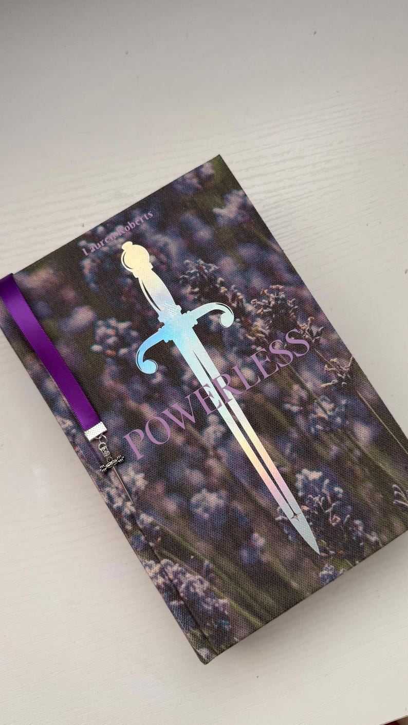 May include: A hardback book titled Powerless by Lauren Roberts featuring a cover image of purple lavender flowers. A large, iridescent, holographic sword graphic is centred on the cover. A purple ribbon bookmark with a small metal sword charm is attached.