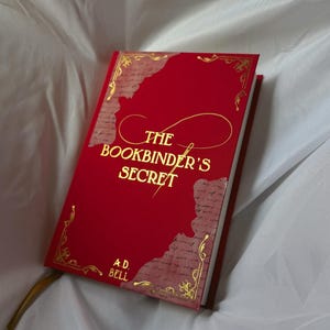 May include: A hardback book titled The Bookbinder's Secret by A.D. Bell with a vibrant red cover, gold foil lettering, and decorative gold corner accents. The cover features a central script design and textured paper-like panels on the sides.