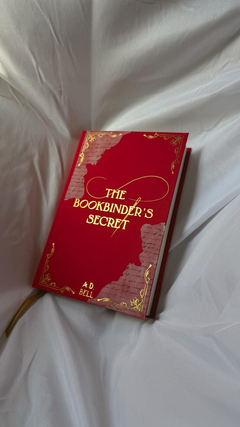 May include: A hardback book titled The Bookbinder's Secret by A.D. Bell with a vibrant red cover, gold foil lettering, and decorative gold corner accents. The cover features a central script design and textured paper-like panels on the sides.