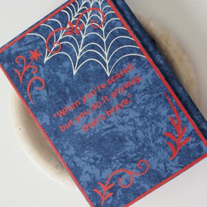 May include: A blue journal with a red border and decorative accents. The cover features a white spiderweb design and the quote, "When you're scared but you do it anyway, that's brave."