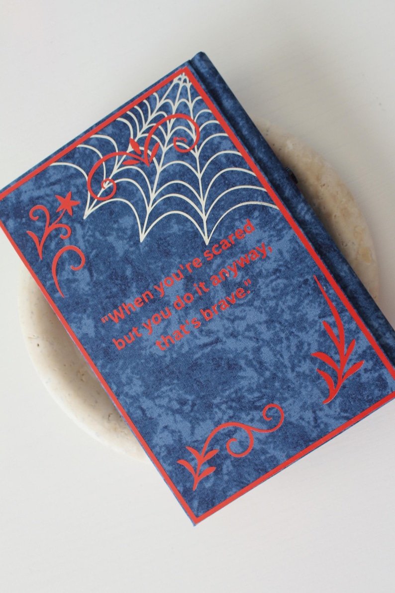May include: A blue journal with a red border and decorative accents. The cover features a white spiderweb design and the quote, "When you're scared but you do it anyway, that's brave."