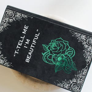 以下が含まれることがあります： 「T-TELL ME I'M BEAUTIFUL」の白文字が入った黒い本。表紙には緑色のバラと蛇のデザイン。白い花のコーナーデザインがテキストと画像を囲んでいます。