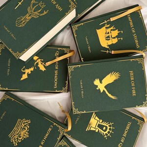 May include: A collection of eight dark green hardback books by Sarah J. Maas, each featuring gold foil illustrations and titles including Kingdom of Ash, Tower of Dawn, Empire of Storms, Queen of Shadows, Heir of Fire, Crown of Midnight, and Throne of Glass.