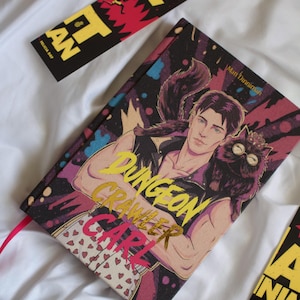 May include: A hardcover book titled Dungeon Crawler Carl featuring an illustration of a man with a cat on his shoulder, wearing heart-patterned boxers. The cover has a dark, textured background with pink and blue accents. Two bookmarks are visible nearby.