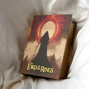 May include: A hardback book titled The Lord of the Rings with a textured cover featuring a dark silhouette of a hooded figure standing before a large, glowing red sun or ring. The title is printed in gold lettering at the bottom of the cover.
