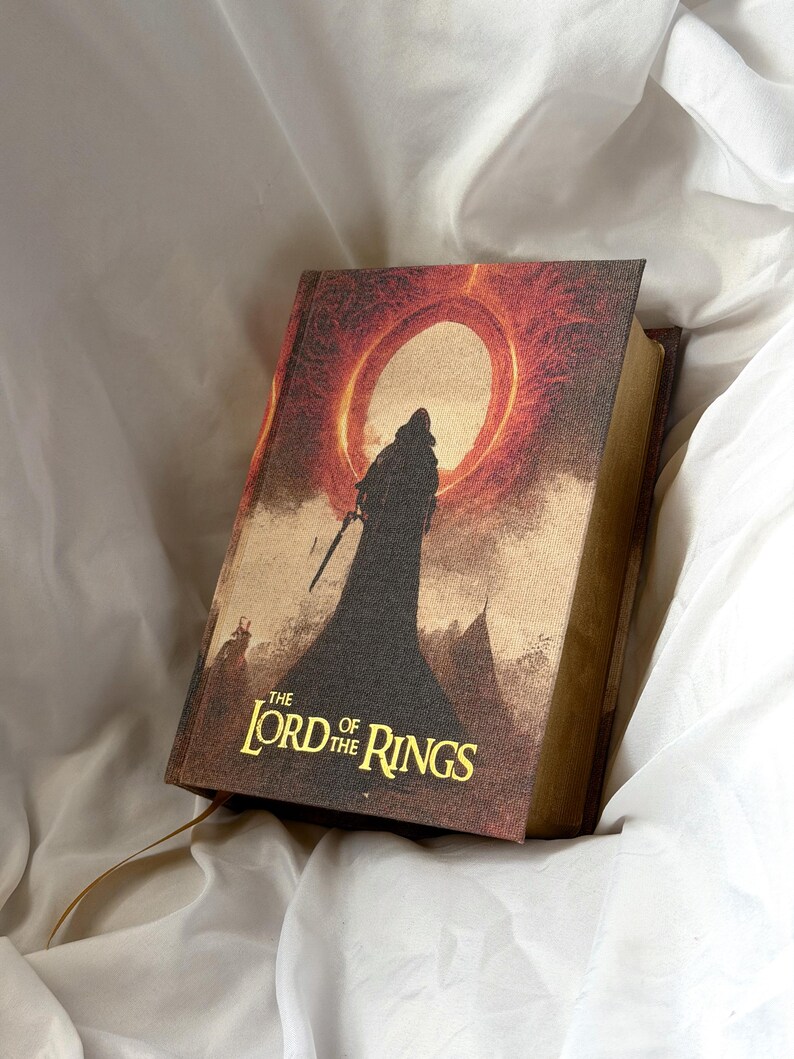 May include: A hardback book titled The Lord of the Rings with a textured cover featuring a dark silhouette of a hooded figure standing before a large, glowing red sun or ring. The title is printed in gold lettering at the bottom of the cover.