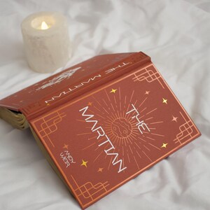 May include: A hardback book titled The Martian by Andy Weir with a rust-coloured cover featuring gold foil celestial graphics, a central planet illustration, and geometric corner borders, positioned next to a lit white pillar candle.