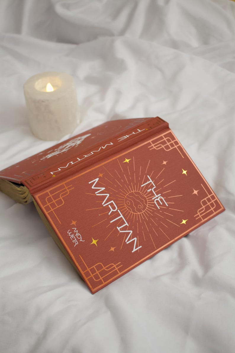 May include: A hardback book titled The Martian by Andy Weir with a rust-coloured cover featuring gold foil celestial graphics, a central planet illustration, and geometric corner borders, positioned next to a lit white pillar candle.