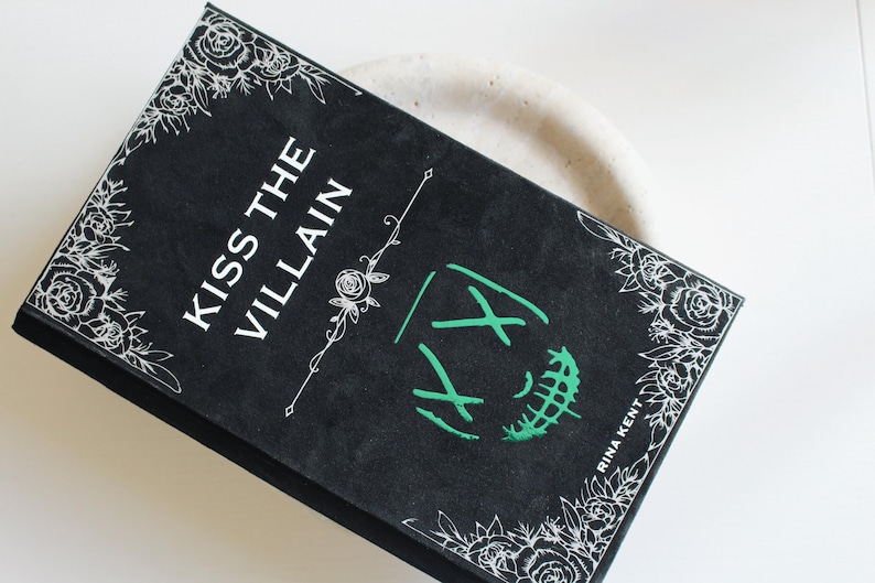 以下が含まれることがあります： 「Kiss the Villain」というタイトルの黒い本。白い文字とコーナーに花の模様があります。表紙には緑色のグラフィック要素と著者の名前が描かれています。本はニュートラルな背景に置かれています。