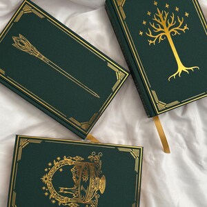 May include: Three dark green hardback books with gold foil detailing. One book features a gold tree and stars, another shows a staff, and the third displays a circular ornate emblem. Each book includes a gold ribbon bookmark and gold corner borders.