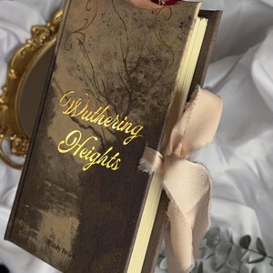May include: A hardback book titled Wuthering Heights by Emily Bront&euml; with a dark brown, textured cover featuring gold foil lettering and decorative flourishes. The book is accented with a light beige, frayed-edge ribbon tied around the side.