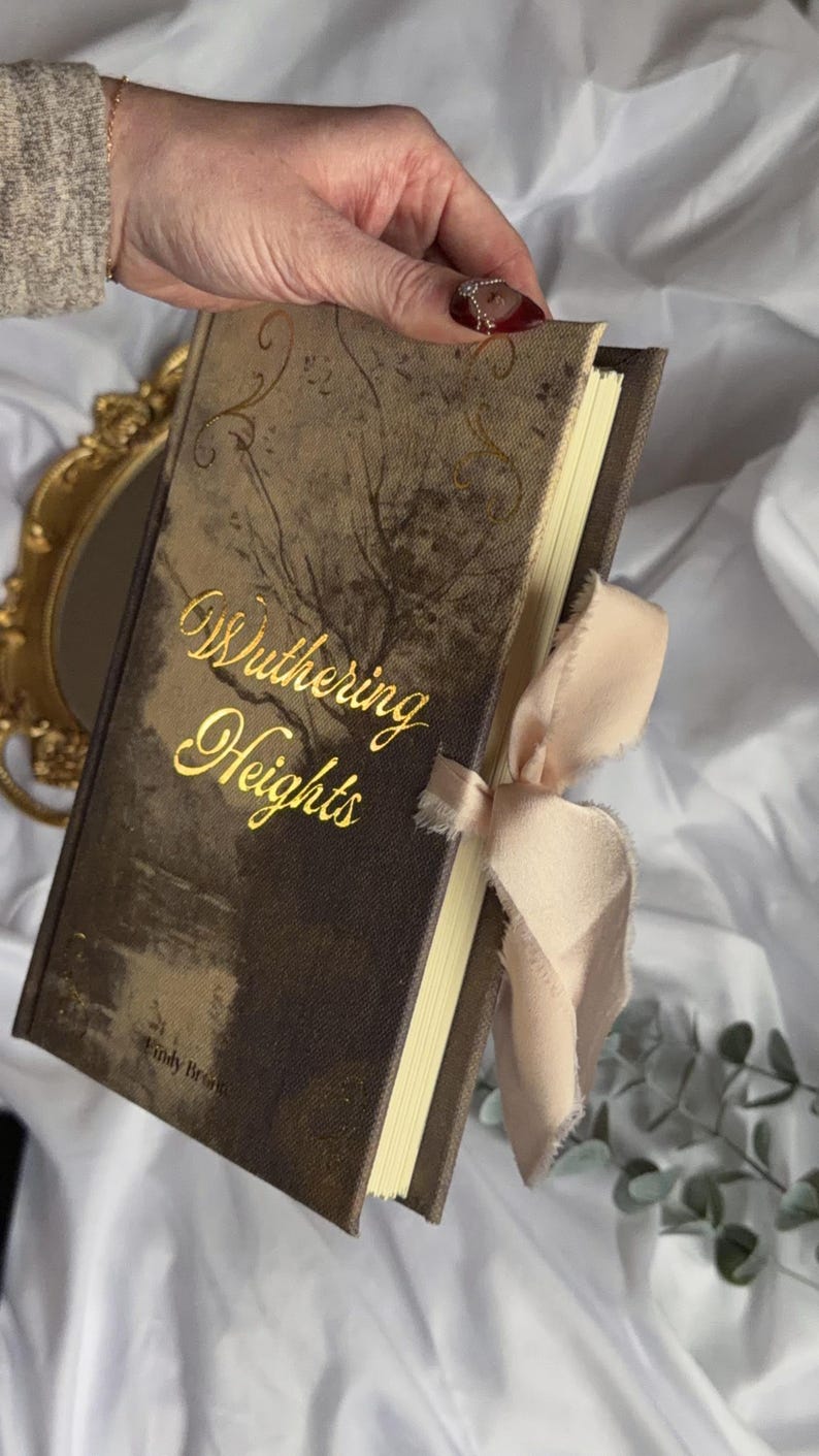 May include: A hardback book titled Wuthering Heights by Emily Bront&euml; with a dark brown, textured cover featuring gold foil lettering and decorative flourishes. The book is accented with a light beige, frayed-edge ribbon tied around the side.
