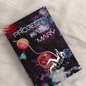 May include: A hardback book titled Project Hail Mary by Andy Weir. The cover features a dark space background with colourful nebulae, planets, and a white silhouette of an astronaut floating while tethered to a curved line across the front.