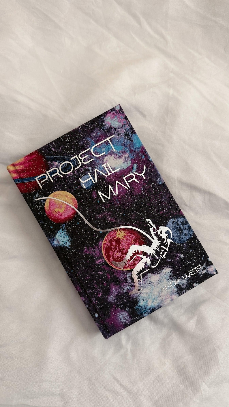 May include: A hardback book titled Project Hail Mary by Andy Weir. The cover features a dark space background with colourful nebulae, planets, and a white silhouette of an astronaut floating while tethered to a curved line across the front.