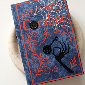 May include: A blue book with the title "Coraline" in orange lettering, featuring a spiderweb design and a black key and hand silhouette. Two black buttons are on the cover, with an orange ribbon bookmark.