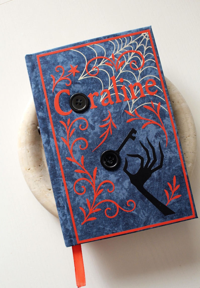 May include: A blue book with the title "Coraline" in orange lettering, featuring a spiderweb design and a black key and hand silhouette. Two black buttons are on the cover, with an orange ribbon bookmark.