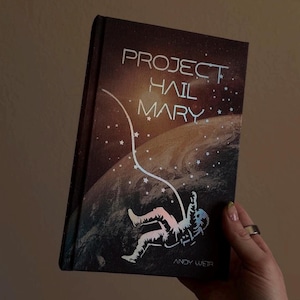 May include: A hardcover book titled "PROJECT HAIL MARY" with an astronaut illustration against a space background. The book cover is predominantly dark brown with white and light blue text and illustrations. The author's name, ANDY WEIR, is at the bottom.