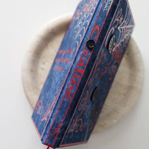 May include: A blue velvet book with red accents and a red ribbon bookmark. The book features a spiderweb design and the word "Charlotte" in red. Two black buttons secure the book's closure. The book is resting on a light-colored, round surface.