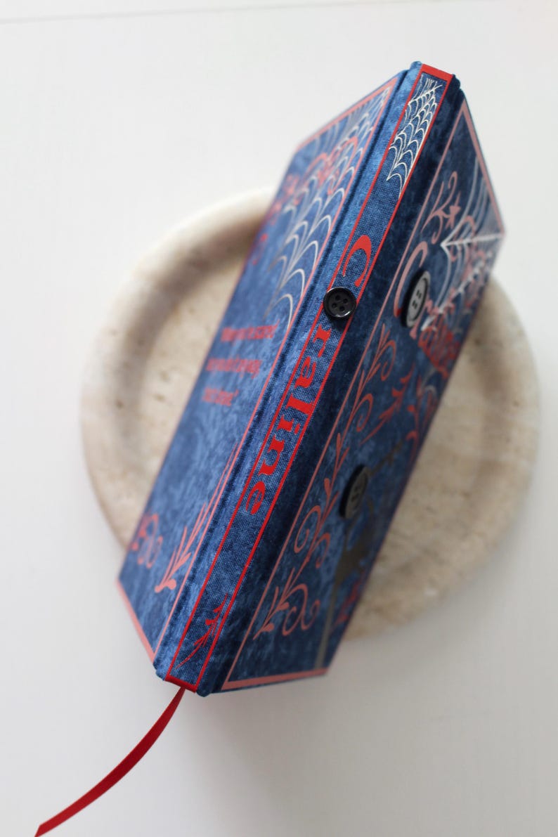 May include: A blue velvet book with red accents and a red ribbon bookmark. The book features a spiderweb design and the word "Charlotte" in red. Two black buttons secure the book's closure. The book is resting on a light-colored, round surface.