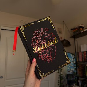 May include: A black hardback book titled Lightlark in gold cursive script. The cover features a pink anatomical heart illustration surrounded by floral elements and gold filigree corner accents. A red ribbon bookmark with a small red bead hangs from the top.