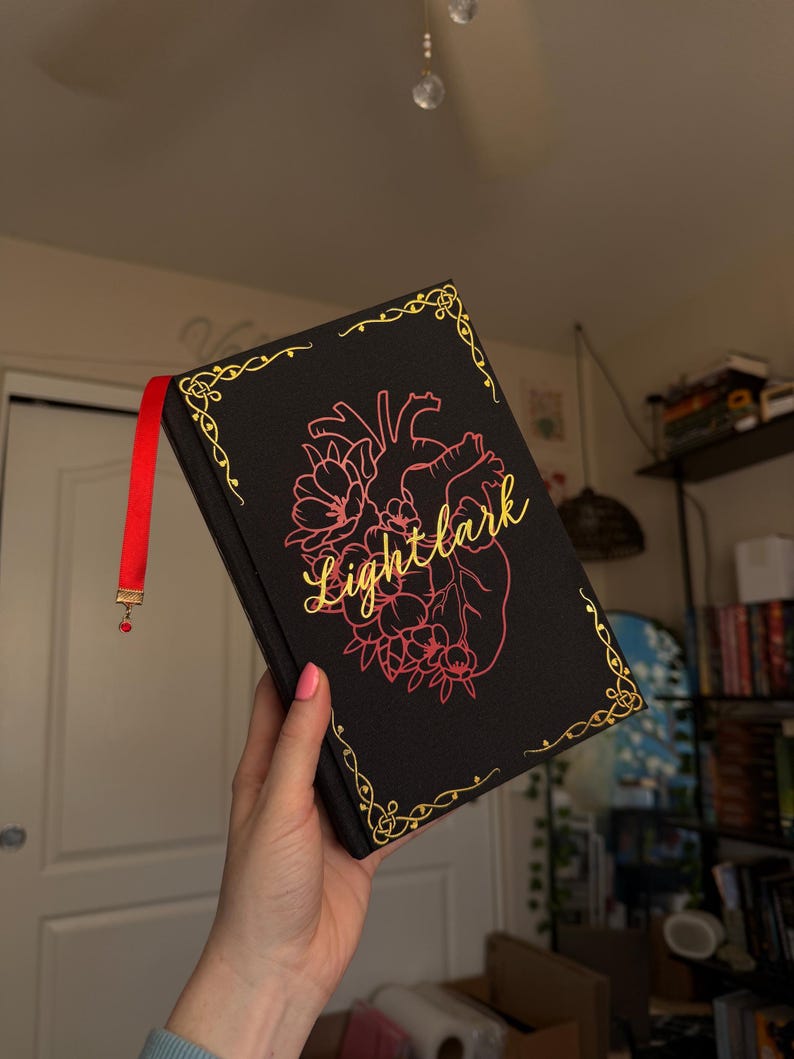 May include: A black hardback book titled Lightlark in gold cursive script. The cover features a pink anatomical heart illustration surrounded by floral elements and gold filigree corner accents. A red ribbon bookmark with a small red bead hangs from the top.