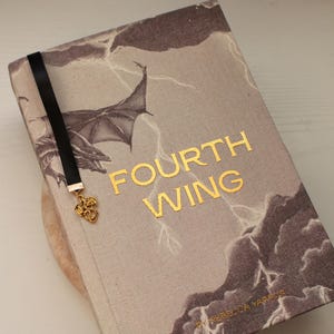 Könnte beinhalten: Ein gebundenes Buch mit dem Titel Fourth Wing von Rebecca Yarros mit einem beigen Stoffeinband, der eine dunkelgraue Illustration von Drachenflügeln und Blitzen zeigt. Ein schwarzes Lesezeichenband mit einem goldenen Drachenanhänger.