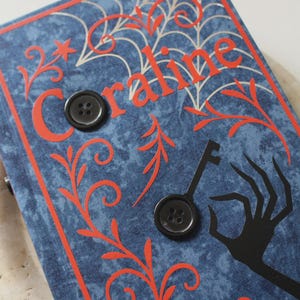 May include: A blue fabric-covered book with the word Coraline in red lettering. The cover features a white spiderweb design, red swirling vine patterns, a black silhouette of a hand holding a key, and two black four-hole buttons attached.
