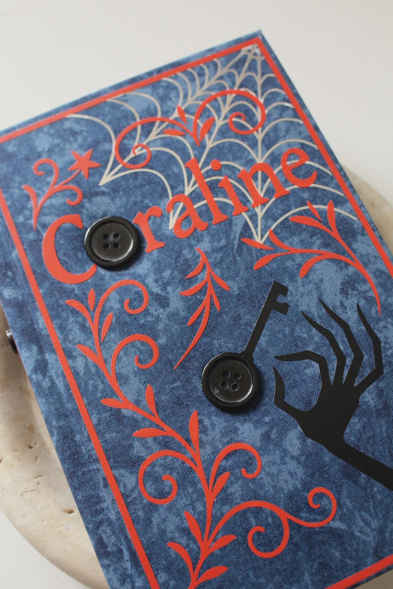 May include: A blue fabric-covered book with the word Coraline in red lettering. The cover features a white spiderweb design, red swirling vine patterns, a black silhouette of a hand holding a key, and two black four-hole buttons attached.